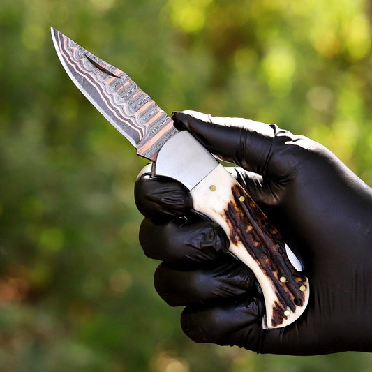 A close-up of a hand wearing a black glove holding a folding knife with a Damascus steel blade and a carved bone handle with dark brown inlays, set against a blurred green outdoor background.