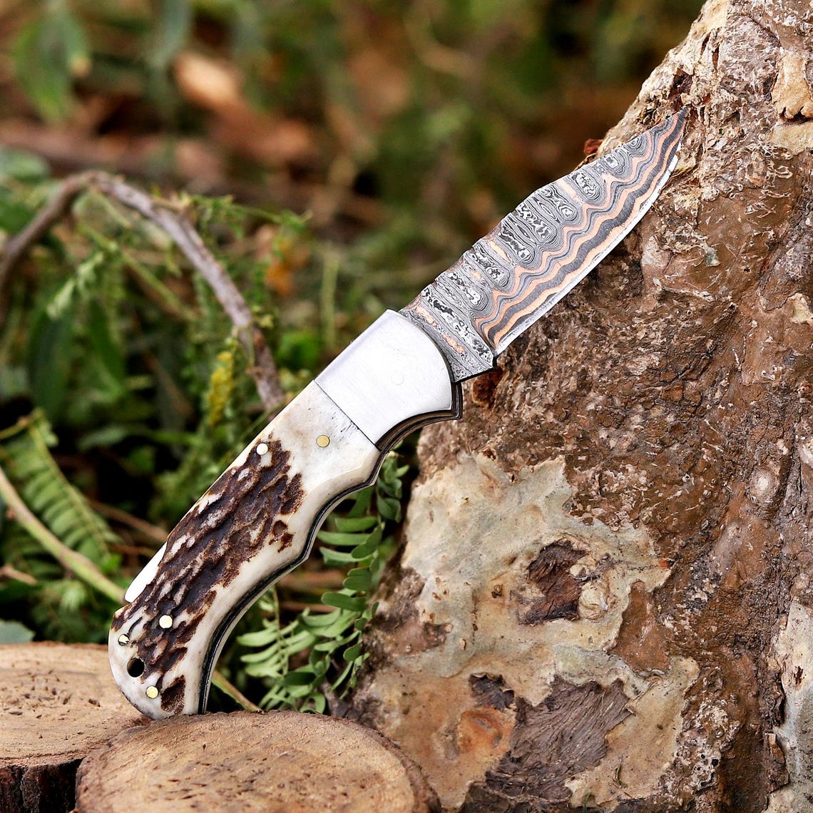 A custom Damascus steel fixed-blade knife with a patterned handle, resting on a tree stump in an outdoor forest setting.