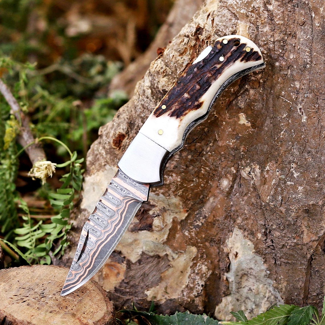 A folding pocket knife with a patterned Damascus steel blade and a bone handle with dark inlay, shown resting on a tree stump outdoors.